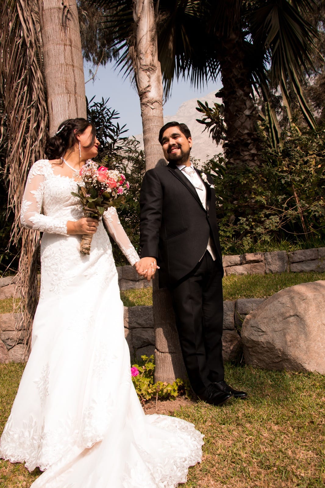 Pareja de novios en traje oscuro y vestido blanco con ramo de flores en una boda al aire libre en Cieneguilla