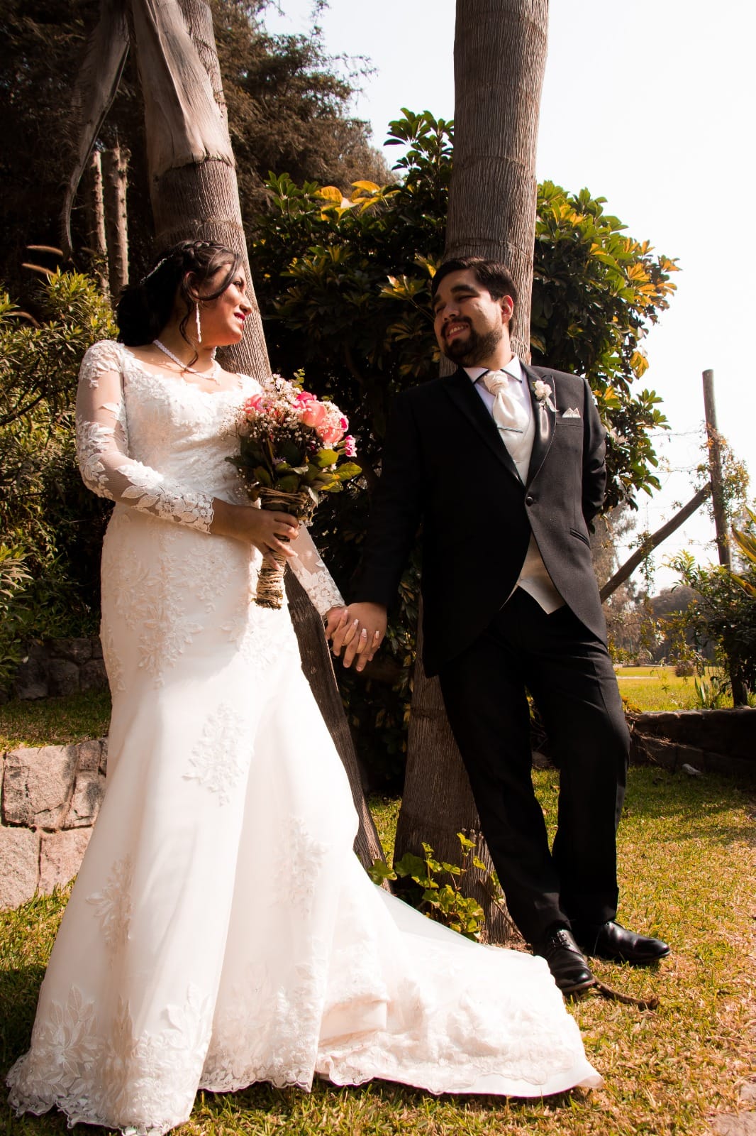Pareja de novios con traje oscuro y vestido blanco sosteniendo un ramo de flores en una boda al aire libre en Cieneguilla