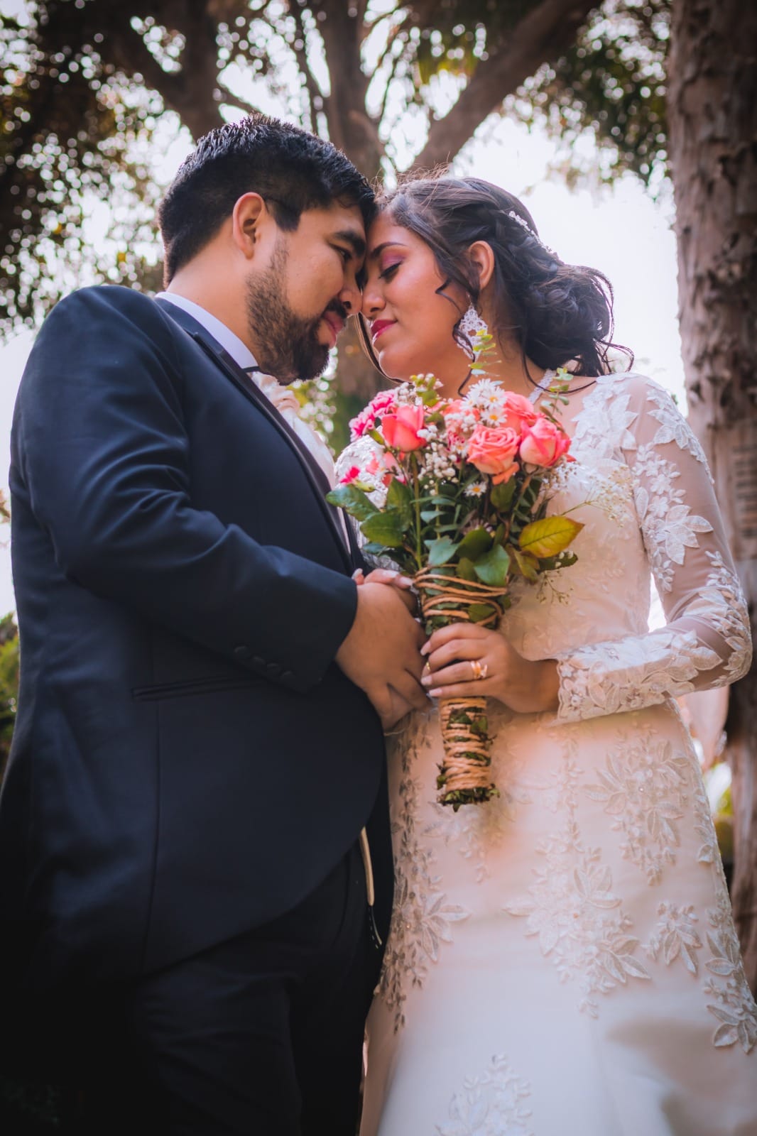 Pareja de novios con traje oscuro y vestido blanco, sosteniendo un ramo de flores en una boda al aire libre en Cieneguilla