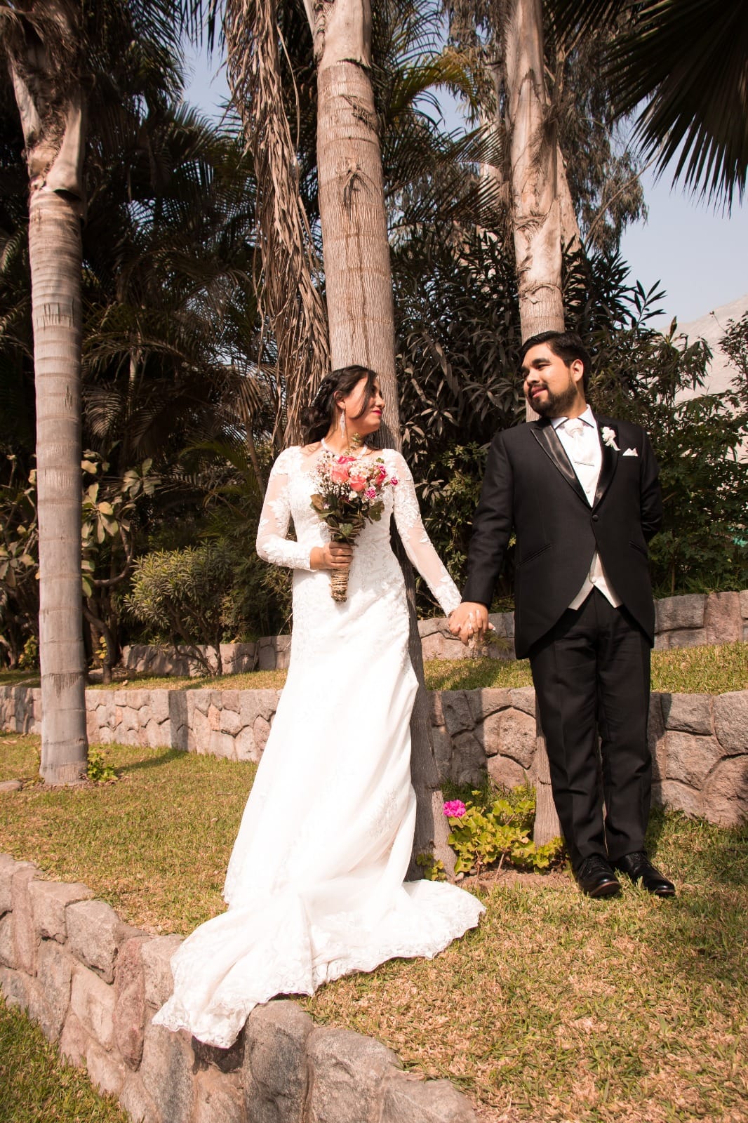 Pareja de novios con traje oscuro y vestido blanco con ramo de flores en una boda al aire libre en Cieneguilla