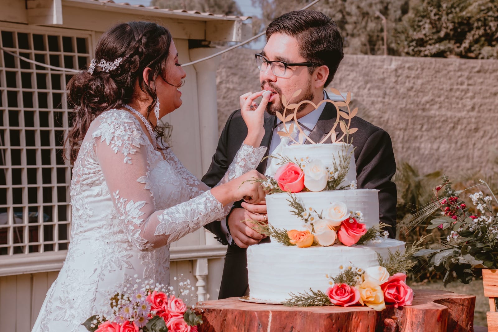 Pareja de novios en traje oscuro y vestido blanco alimentándose con pastel de bodas en Cieneguilla
