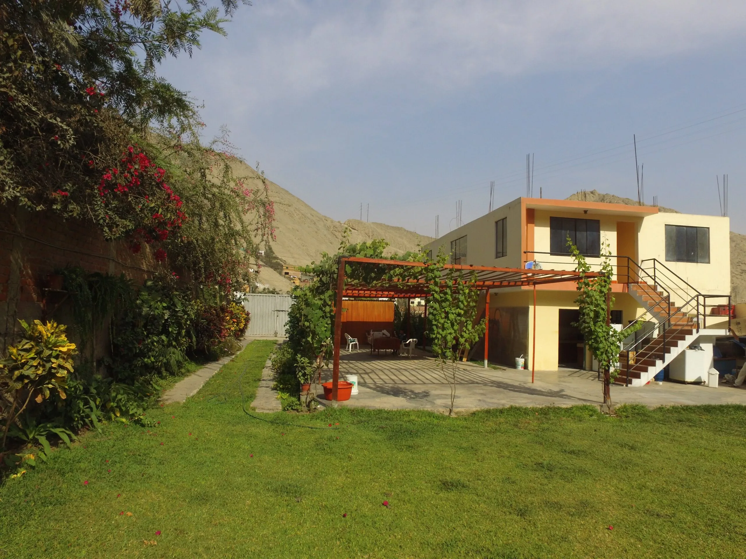 Casa de dos pisos con pérgola en el patio, rodeada de plantas y flores, en Cieneguilla