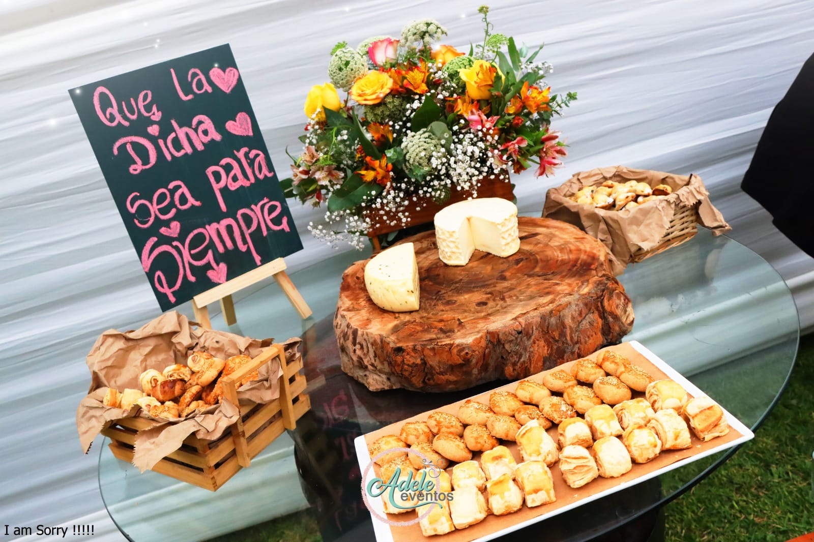 Variedad de alimentos en una mesa de cristal, incluyendo panes y pasteles en una cesta de madera y una bandeja, junto con un tronco de madera con queso, flores coloridas en la decoración y una pizarra con la frase 'Que la dicha sea para siempre'.
