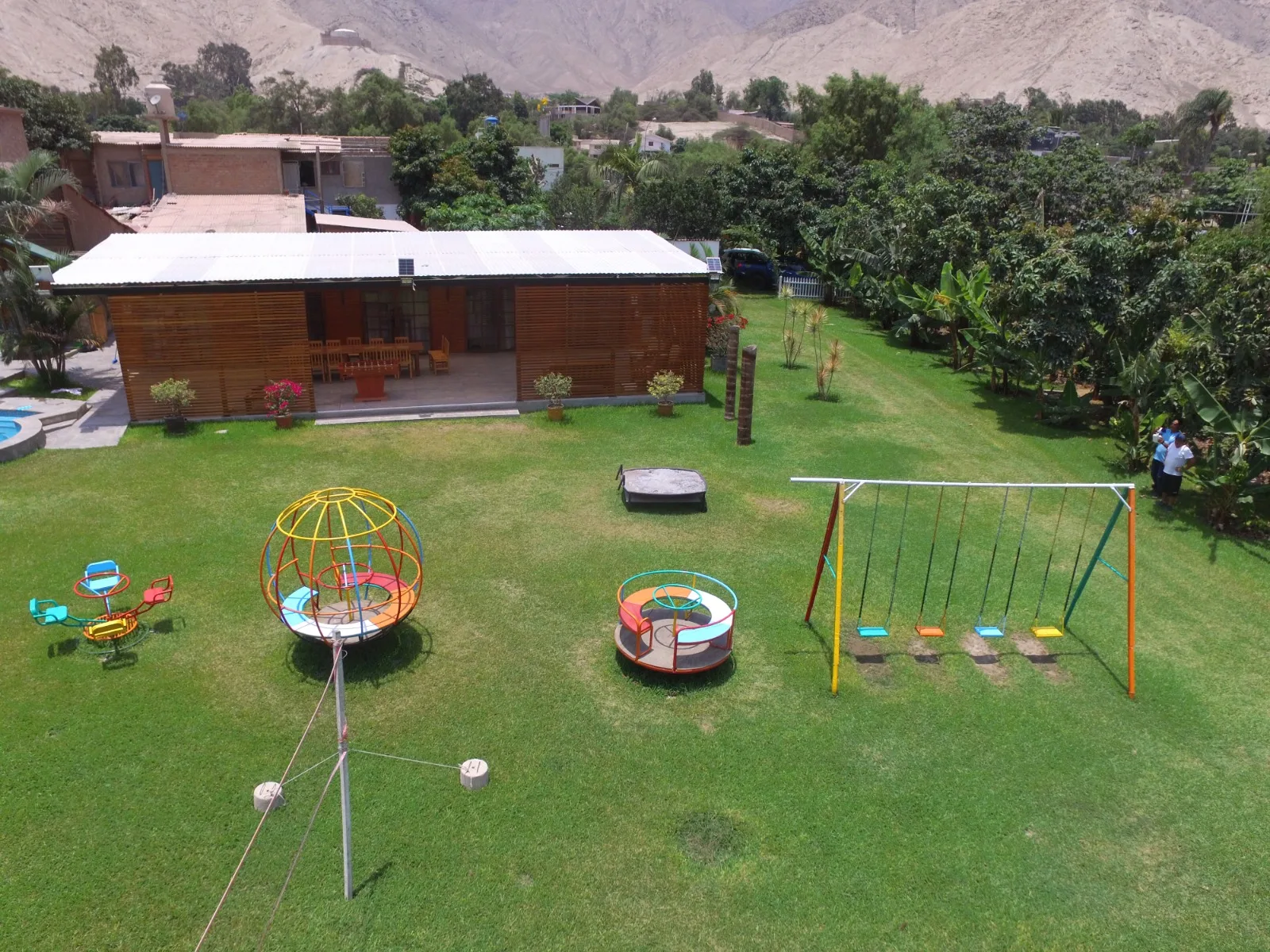 Área de juegos infantiles con césped, estructuras de juego coloridas y un edificio con terraza cubierta en una casa en alquiler en Cieneguilla.