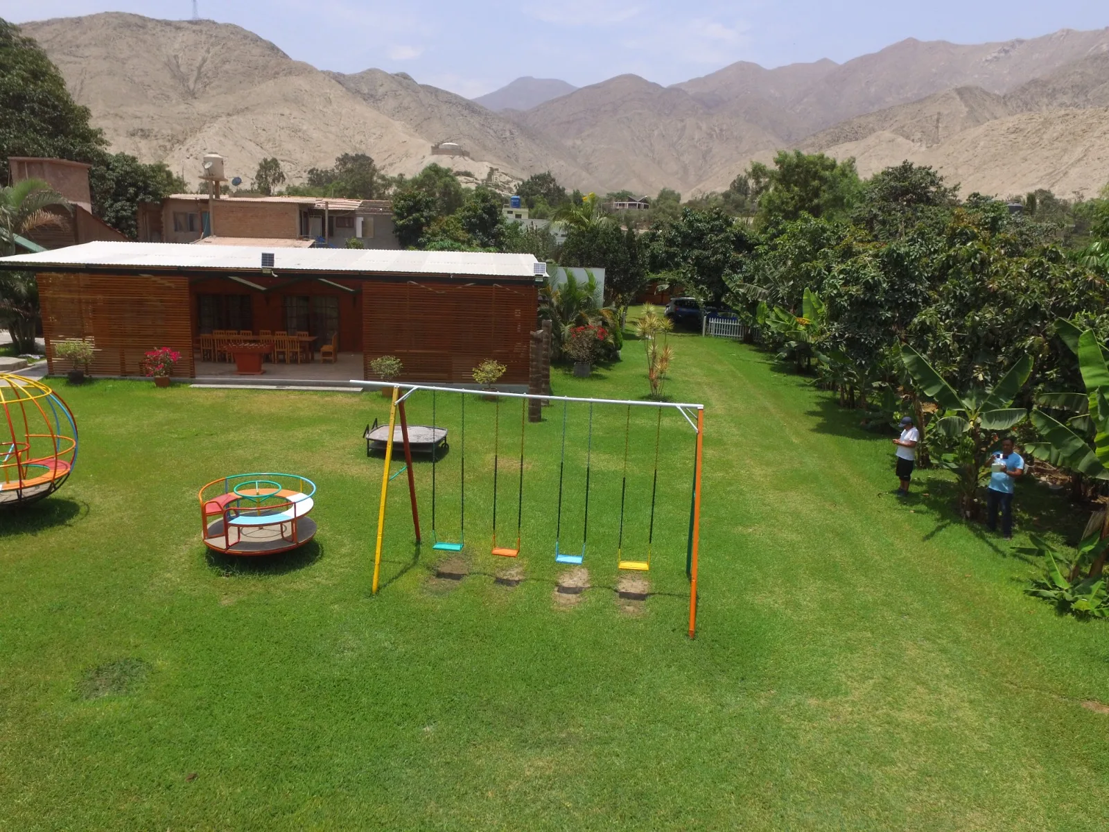 Área de juegos infantiles con césped, estructuras de juego coloridas y un edificio de madera con terraza cubierta en una casa en alquiler en Cieneguilla.