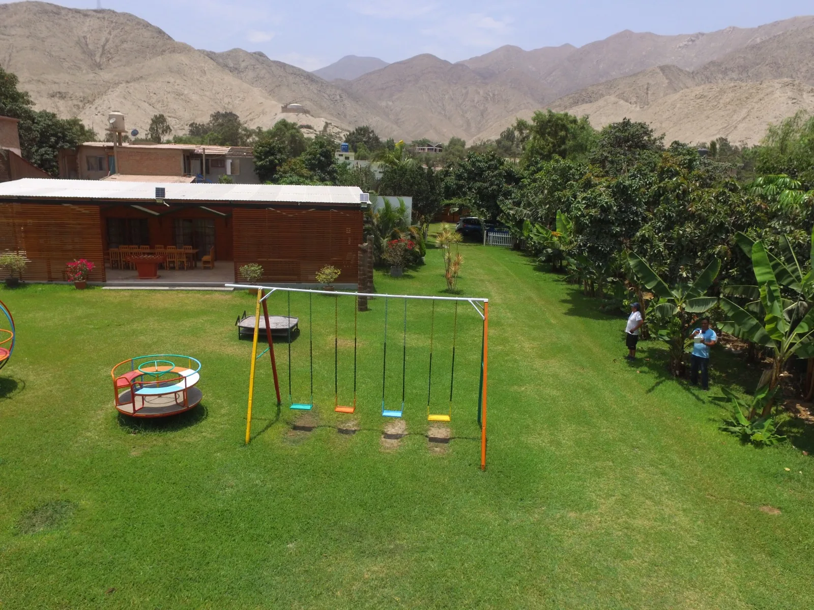 Área de juegos infantiles con columpios y carrusel en un jardín con césped. Al fondo, hay una terraza cubierta con sillas y mesas, rodeada de árboles y plantas con montañas en la distancia.