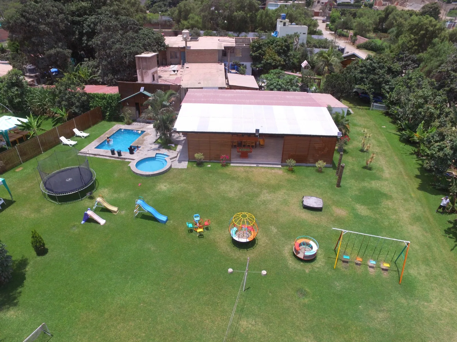 Vista aérea de un área recreativa con piscina, jacuzzi, césped, trampolín, área de juegos infantiles y mobiliario de exterior en una casa en alquiler en Cieneguilla.