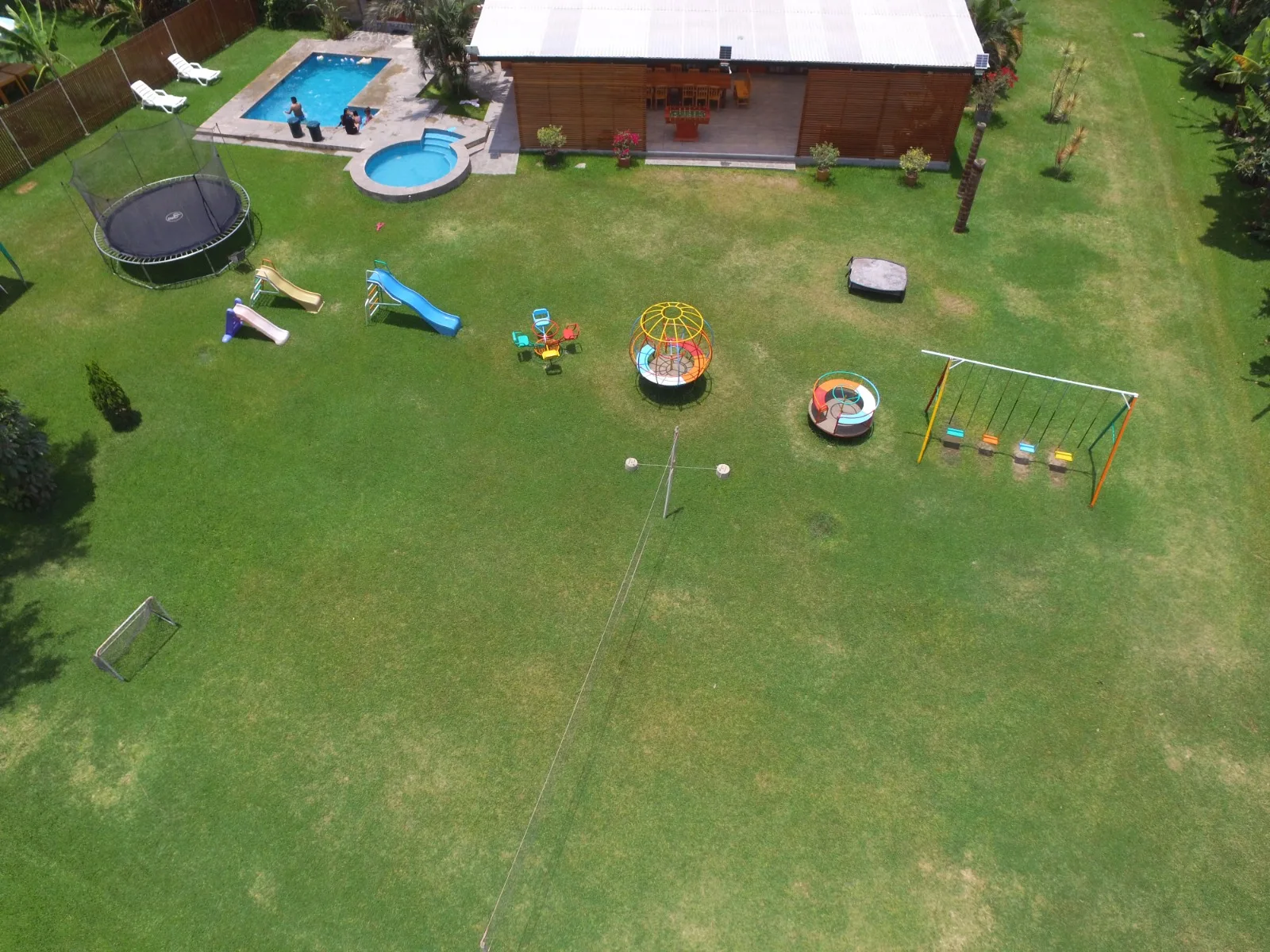 Vista aérea de un jardín trasero con piscina, jacuzzi, césped, trampolín, área de juegos infantiles y mobiliario de exterior en una casa en alquiler en Cieneguilla.