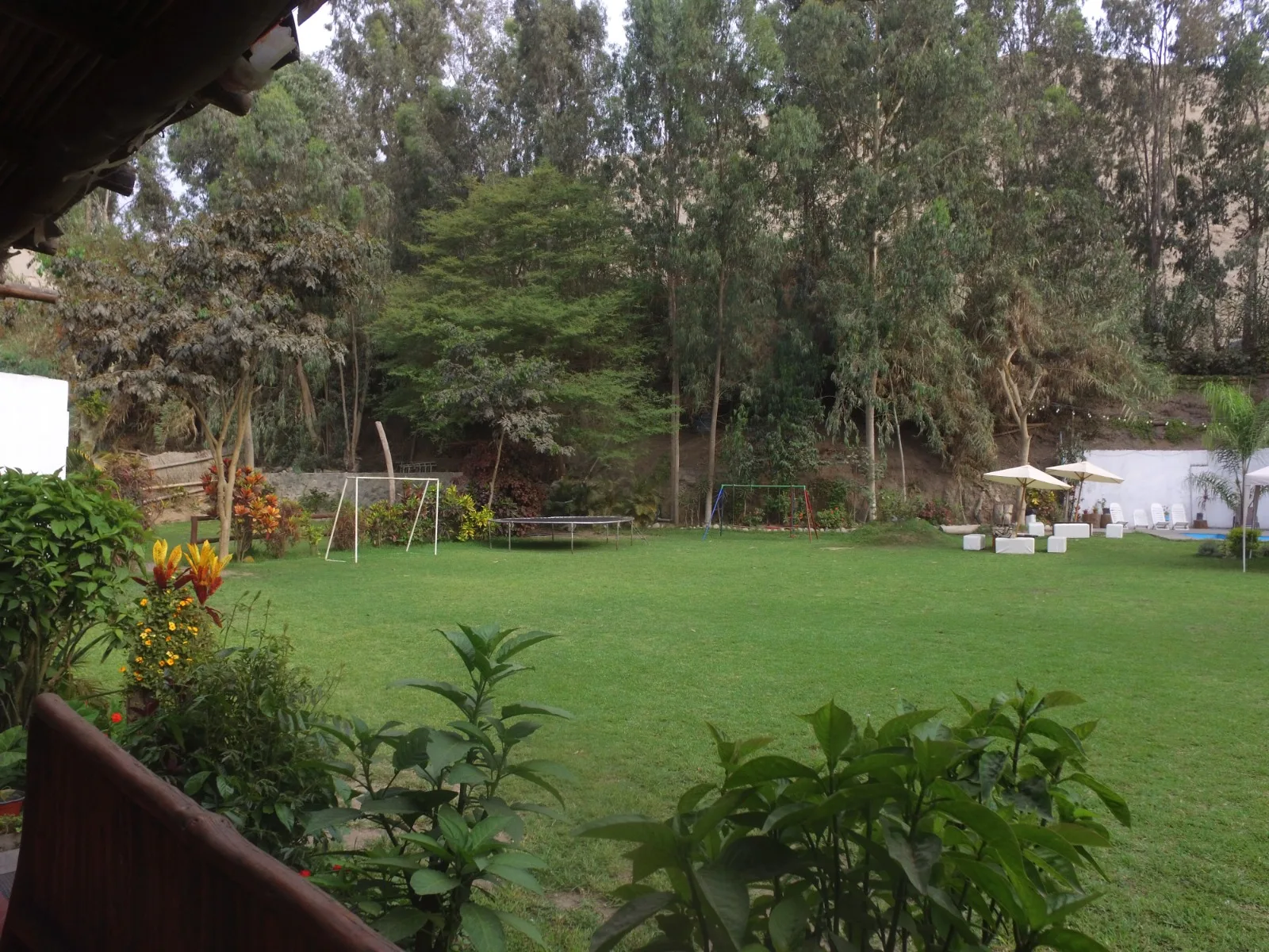 Jardín trasero con césped, columpios, una cama elástica y una zona de descanso con mesas y sillas bajo sombrillas blancas en una casa en alquiler en Cieneguilla.