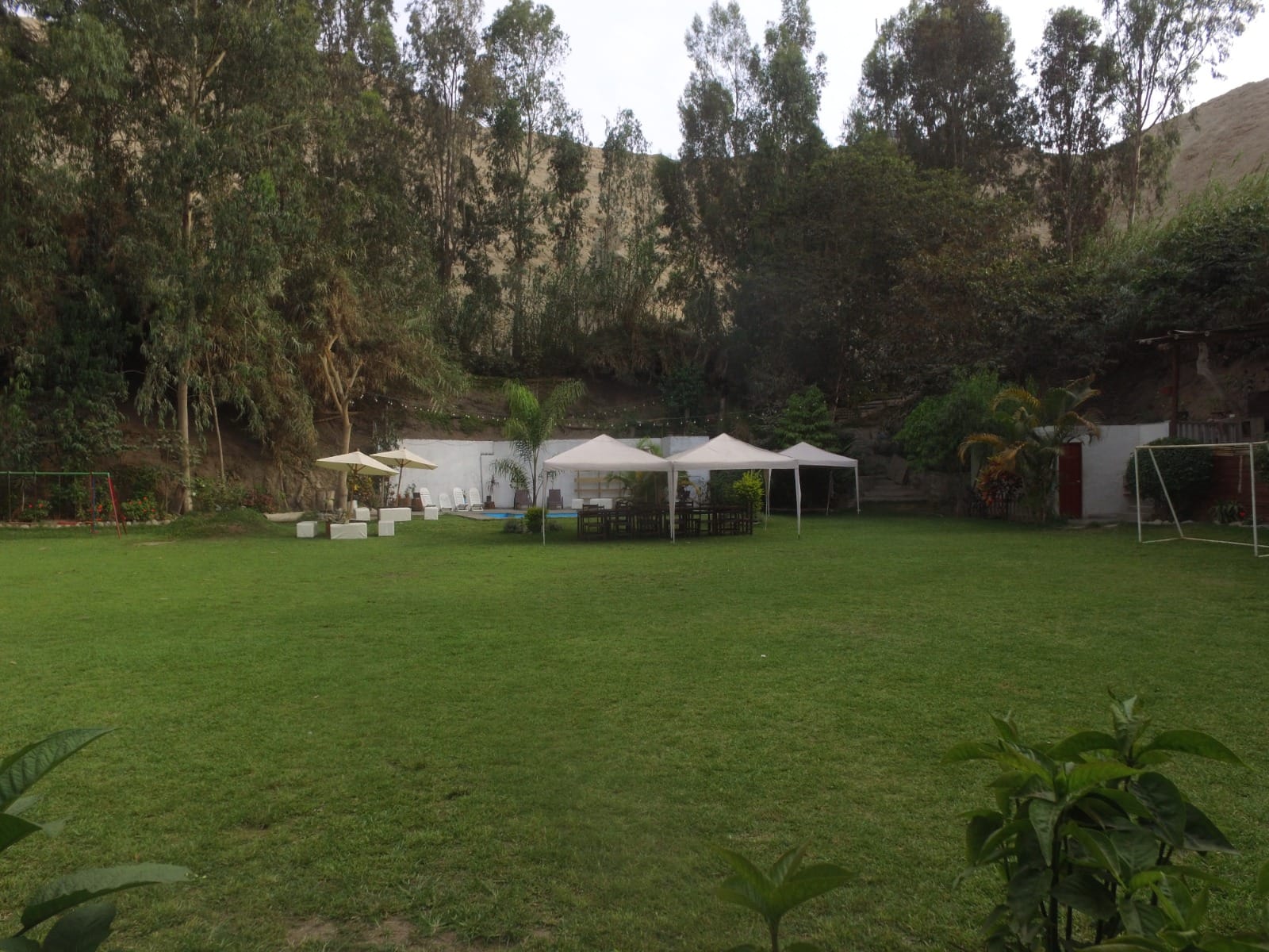 Patio exterior con césped, toldos blancos, sillas y mesas, columpios y una portería de fútbol en una casa en alquiler en Cieneguilla.