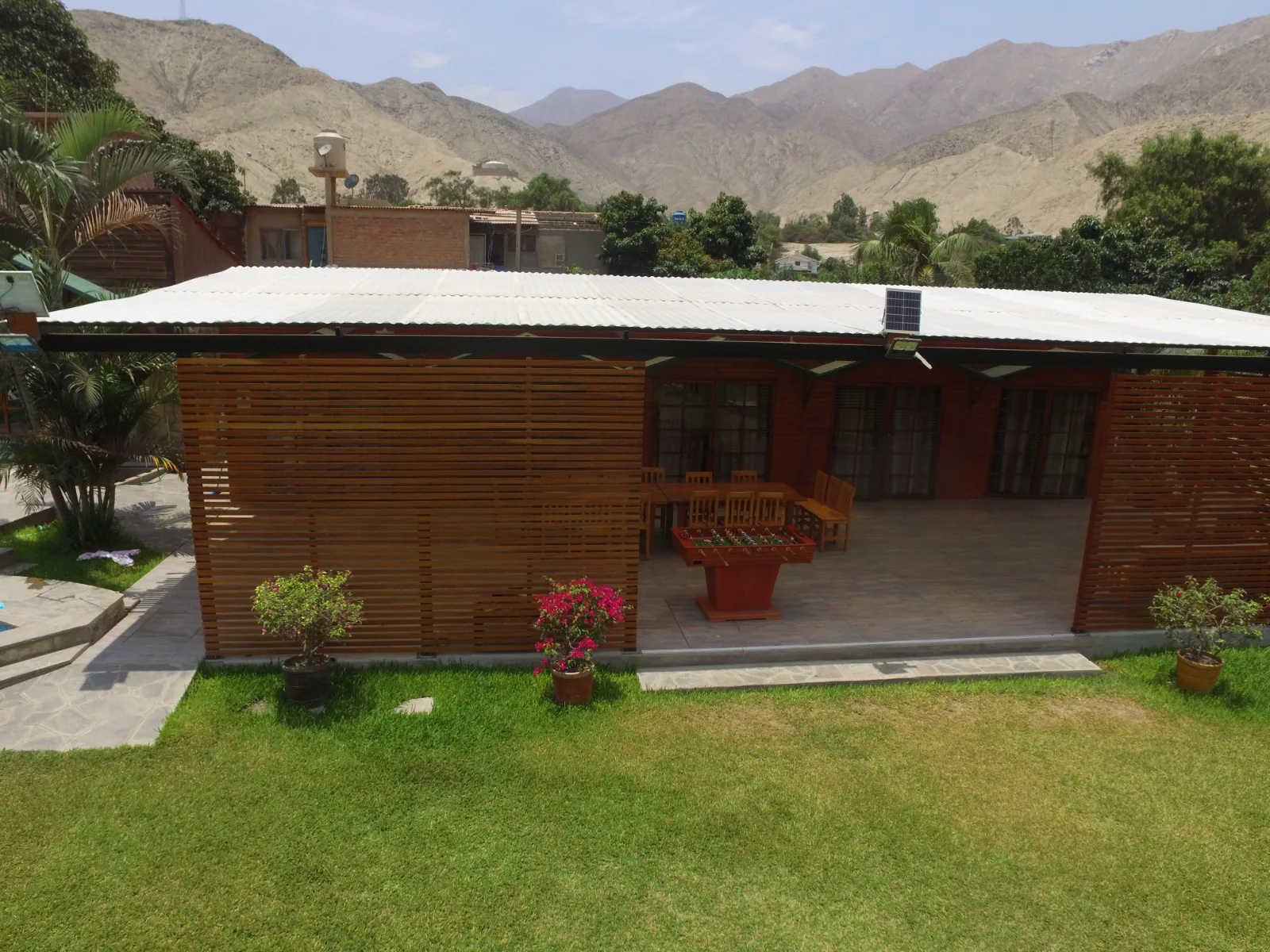 Edificio de una sola planta con techo de metal corrugado y paredes de listones de madera, con un patio cubierto que tiene varias sillas de madera y una mesa, situado en una zona de césped con un camino de concreto que lleva al patio.