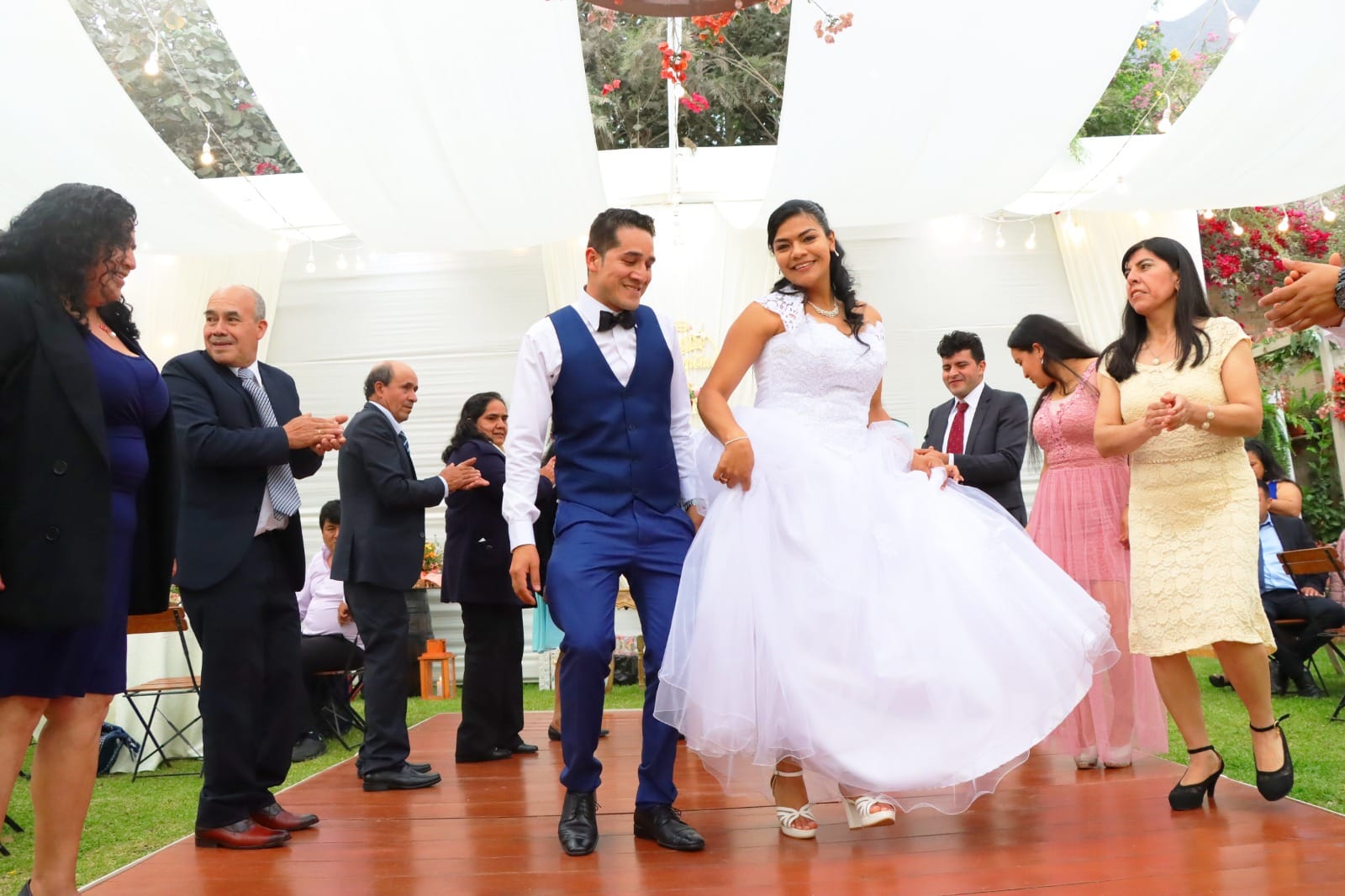 Pareja de novios bailando en una plataforma de madera durante su boda en Cieneguilla, con la novia en un vestido blanco y el novio en un traje azul, rodeados de invitados celebrando al aire libre bajo un canopy decorado.