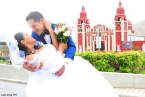 Pareja de novios posando afuera de una iglesia en Cieneguilla, con la novia en un vestido blanco y el novio en un traje azul.