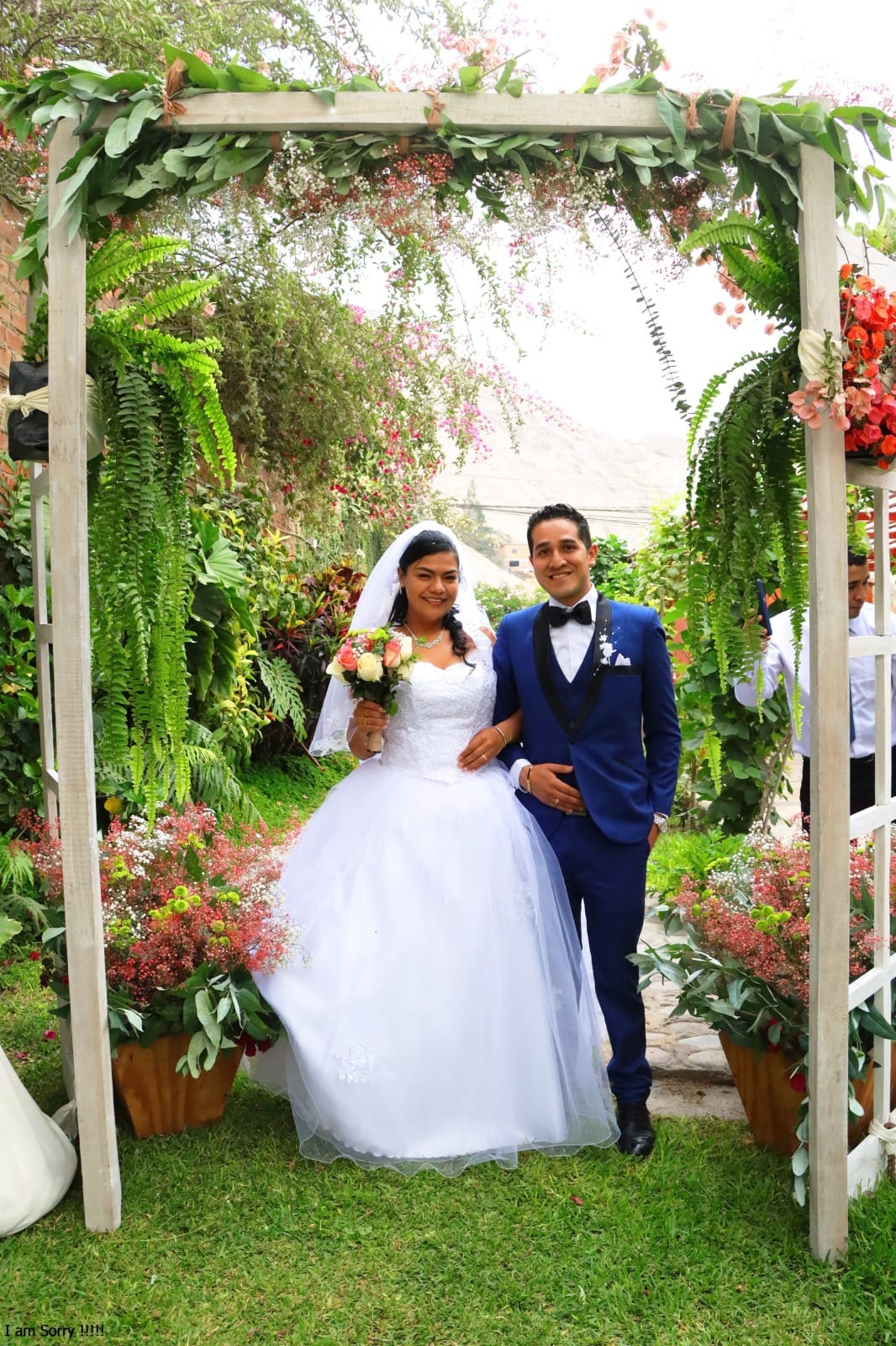 Pareja de novios bajo un arco decorado con follaje verde y flores durante su boda al aire libre en Cieneguilla, con la novia en un vestido blanco y el novio en un traje azul.