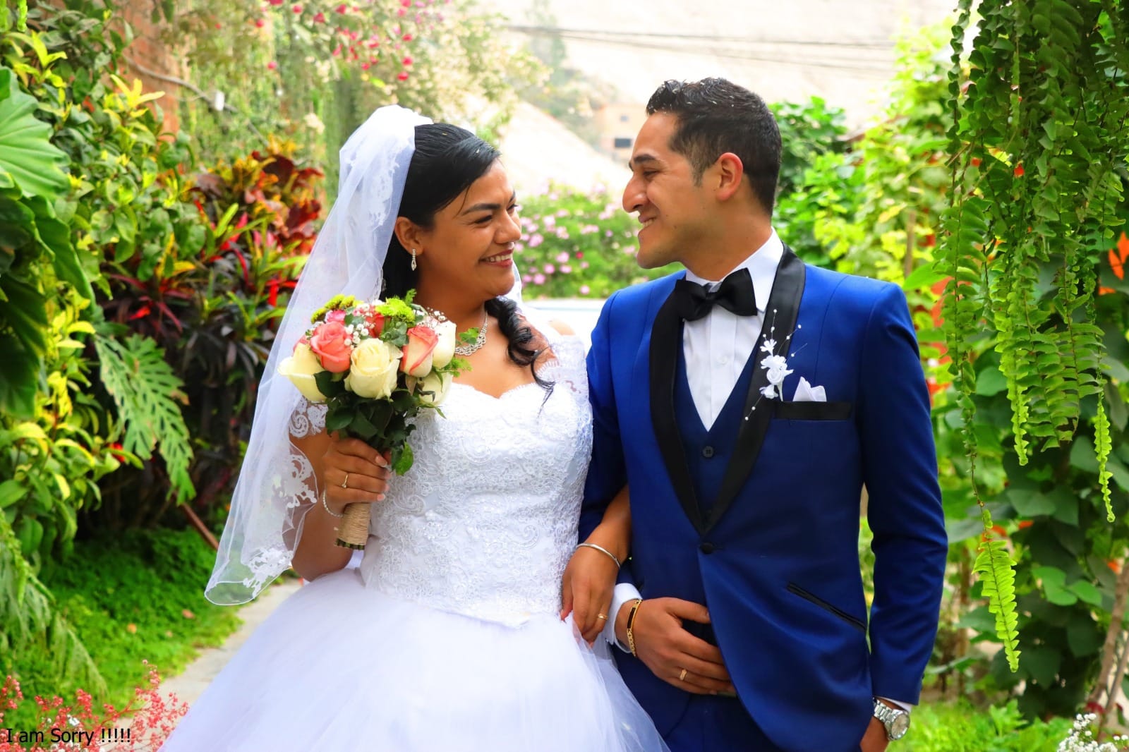 Novia con vestido blanco y velo sosteniendo un ramo de flores, junto con el novio en traje azul con pajarita negra, posando en un jardín durante su boda en Cieneguilla.