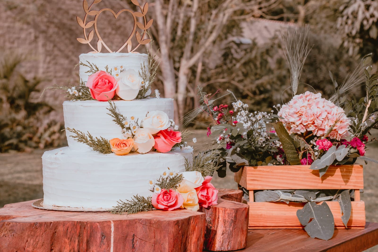 Pastel de boda de tres niveles decorado con flores, incluyendo rosas rosas, blancas y naranjas, y flores pequeñas blancas, junto con follaje verde. El pastel está coronado con un topper de madera en forma de corazón con hojas. Está colocado sobre una losa de madera junto a una caja de madera llena de flores variadas. El fondo muestra un entorno al aire libre con árboles y plantas.