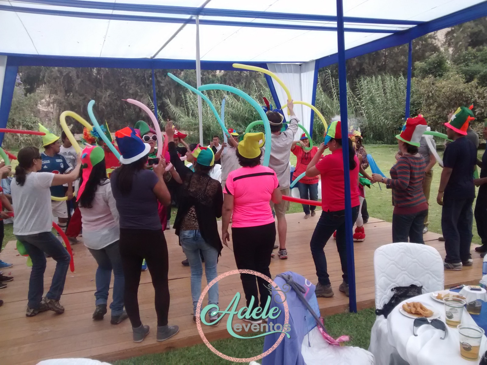Colaboradores de una empresa bailando con sombreros festivos y globos bajo un toldo durante un evento de Adele Eventos en Cieneguilla.